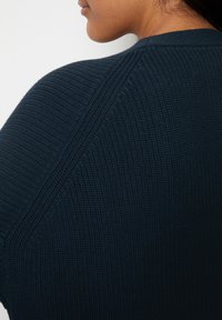 Persoon die een donkerblauw geribd gebreide trui draagt, met de achterkant en zijkant van de nek- en schouderpartij zichtbaar.