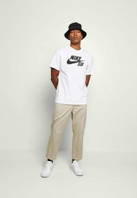 Mann iført svart bøttehatt, hvit Nike SB t-skjorte, beige bukser, svarte sokker og hvite sneakers, stående mot en ensfarget lys vegg.