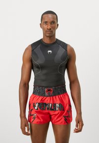 Man wearing black sleeveless compression shirt and red Venum boxing shorts, standing against a plain light background.