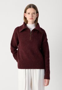 Bordeaux gebreide pullover met een kwart-rits kraag, ribbelboorden en gestructureerde textuur. Draagt over een witte coltrui en gecombineerd met lichte broeken.
