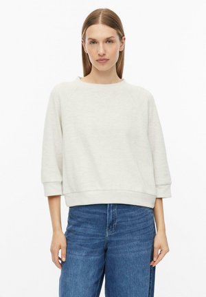 Frau mit glatten braunen Haaren trägt ein lockeres weißes Langarmshirt und blaue Jeans, steht mit neutralem Gesichtsausdruck vor weißem Hintergrund.