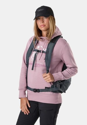 Junge Frau mit pinkem Hoodie, schwarzer Kappe und Rucksack mit Brust- und Hüftgurt, die vor einem schlichten hellen Hintergrund posiert.