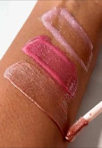 Trois échantillons de gloss à lèvres sur la peau : rose clair avec des paillettes, rose vif et rose nude doux avec des paillettes, appliqués avec un pinceau à lèvres.