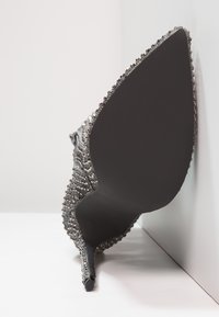 Chaussure à talons hauts avec une semelle noire, un tissu gris orné de perles argentées, un bout pointu et des détails texturés le long du bord supérieur.