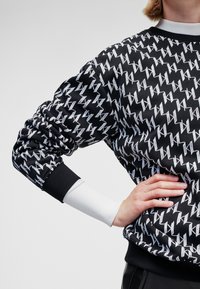 Schwarzer Pullover mit auffälligem weißem Zickzackmuster. Gerippte Bündchen und Kragen, mit strukturiertem Stoff und lockerer Passform an den Ärmeln.