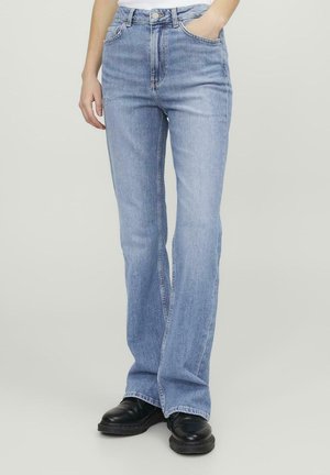 Persona che indossa jeans a gamba dritta azzurri chiaro con scarpe nere, una mano in tasca, in piedi contro uno sfondo chiaro uniforme.