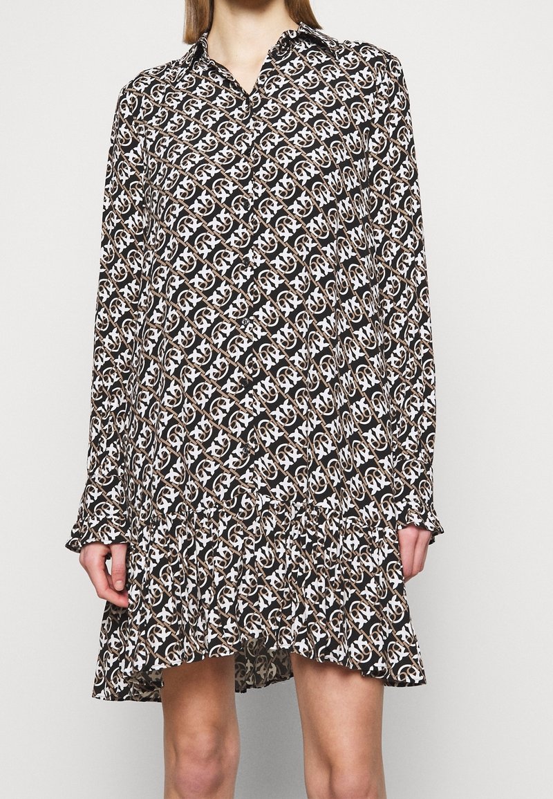 Vrouw draagt een jurk met lange mouwen en knopen, gemaakt van stof met een abstract kettingpatroon in zwart, wit en bruin, en een gerimpelde zoom.