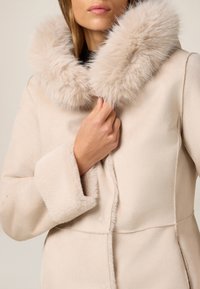 Cappotto beige chiaro con colletto in pelliccia chiara e morbida. Tessuto soffice, design strutturato e chiusura con bottoni. Mano posizionata sul colletto.