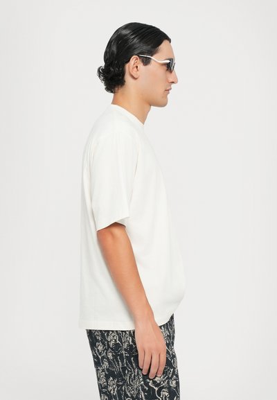 Tricou alb cu mânecă scurtă și guler rotund, asortat cu pantaloni închisi la culoare cu un model. Modelul poartă ochelari de soare eleganți și are părul aranjat cu gel.