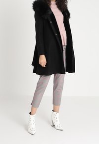 Schwarzer Wollmantel mit Kapuze aus Kunstfell, kombiniert mit einem rosa Rippoberteil, karierten Hosen mit blauen und rosa Längsstreifen und weißen Ankle Boots mit Ösenverzierung.