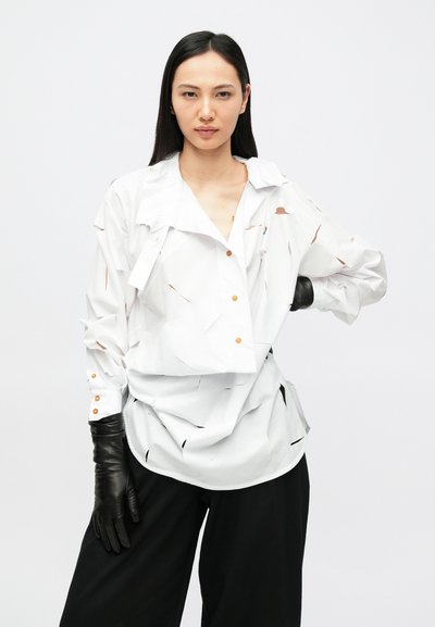 Λευκό πουκάμισο με σχέδια κοπής, πορτοκαλί κουμπιά και διπλωμένο γιακά. Συνδυάζεται με μαύρα δερμάτινα γάντια και φαρδιά μαύρα παντελόνια.