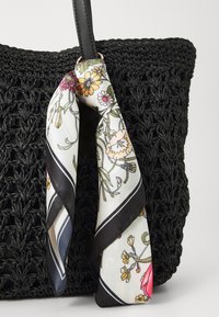 Sac cabas tissé noir avec une finition texturée. Un foulard en soie à motif floral est attaché à une des lanières, présentant des couleurs vives et des accents noirs.