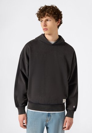 Giovane uomo con capelli ricci che indossa una felpa nera con cappuccio e jeans azzurri chiari, guarda verso sinistra su uno sfondo bianco.