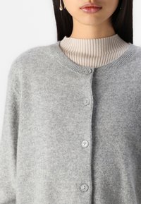 Femme portant un cardigan en laine gris clair boutonné sur un haut beige côtelé à col montant, montrant les lèvres et le bas du visage avec des cheveux raides et foncés.