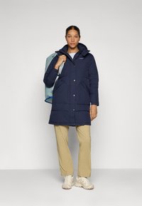 Marineblauwe geïsoleerde parka met een capuchon, voorzien van voorzakken, een knoopsluiting, en gedragen over beige broeken en lichte sneakers.