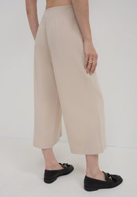 Person in beigen, weit geschnittenen, knöchellangen Hosen, schwarzen Loafers mit Quasten und einem goldenen Ring, steht auf einem weißen Boden vor einer schlichten Wand.