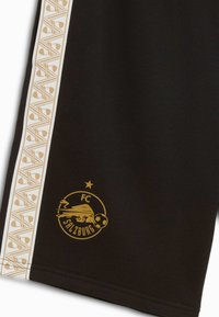 Pantalones cortos deportivos negros con un logo dorado del FC Salzburg. Las franjas laterales blancas con patrones geométricos realzan el diseño. Textura de tela suave.