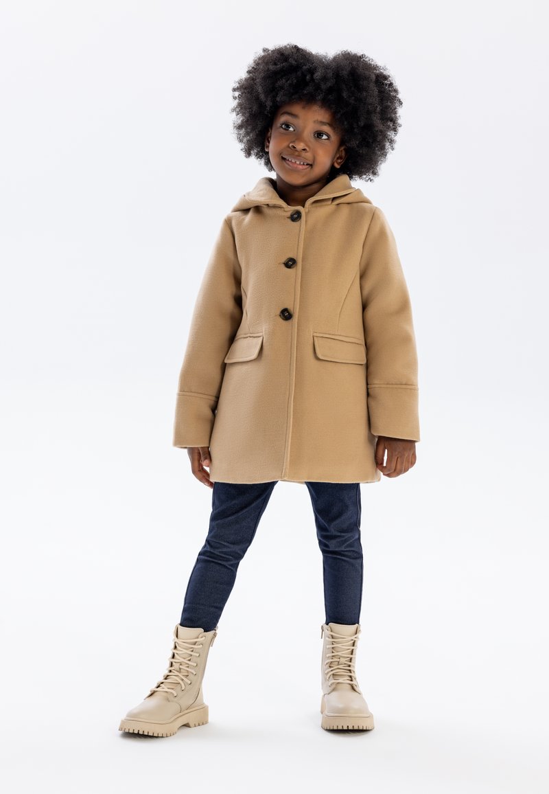 Manteau à capuche beige avec des boutons à l'avant et deux poches, associé à un jean bleu foncé et des bottes beiges à lacets. Texture lisse.