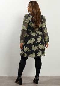 Vestido negro floral con tejido transparente, mangas largas y acentos metálicos en dorado. Presenta una cintura ajustada y una falda acampanada, combinado con medias negras.