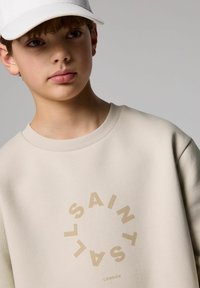 Bež majica s kapuco z okroglim bež tiskom s besedilom "SAINT" in "LONDON" spodaj. Mehak material s okroglim izrezom in rebrastimi manšeti.