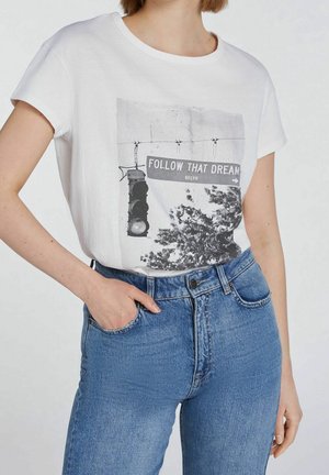 Femme portant un t-shirt blanc avec un feu de circulation et un panneau indiquant "Follow That Dream BKLYN", associé à un jean bleu taille haute.