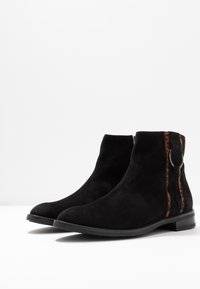 Stivali alla caviglia in suede nero con una finitura liscia, dotati di una zip laterale e una striscia di accentuazione marrone con un dettaglio hardware circolare. Suola piatta.