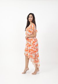 Asymmetrisches Kleid in Orange mit Blumenmustern, kurzen Rüschensäumen und einer taillierten Taille. Kombiniert mit transparenten Absatzsandalen.