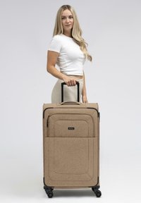 Grande valigia con ruote in tessuto beige, caratterizzata da una forma rettangolare aerodinamica, dettagli neri e una robusta maniglia retrattile.