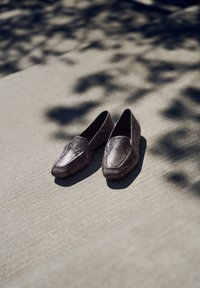 Paire de mocassins marron foncé à motif peau de serpent posée sur un béton ensoleillé avec des ombres douces d’arbres proches.