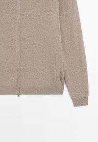 Pull zippé beige en maille, manches longues, poignets et ourlet côtelés, avec poches latérales sur fond blanc.