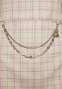 Beżowa tkanina w kratę z fioletowymi i szarymi liniami, z dwiema nałożonymi srebrnymi łańcuszkami przyczepionymi za pomocą dekoracyjnych zawieszek.