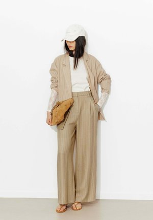 Femme en tailleur beige à fines rayures avec manches en dentelle blanche, casquette blanche, sac en daim marron clair et sandales, debout contre un mur blanc.