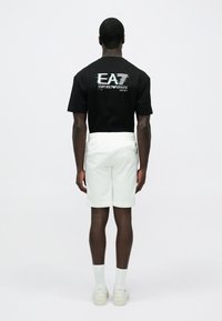 Vīrietis stāv ar muguru pret skatītāju, valkājot melnu EA7 Emporio Armani T-kreklu, baltus šortus, baltas zeķes un baltas sporta kurpes uz vienkārša fona.