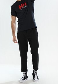 Man som bär en svart Helly Hansen T-shirt, svarta korta byxor och svarta högstövlar, stående mot en vit bakgrund.