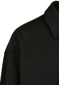 Musta lyhythihainen paita, jossa on laskostettava kaulus, sileä pinta ja rento malli, korostaen minimalistisia muotoiluyksityiskohtia.