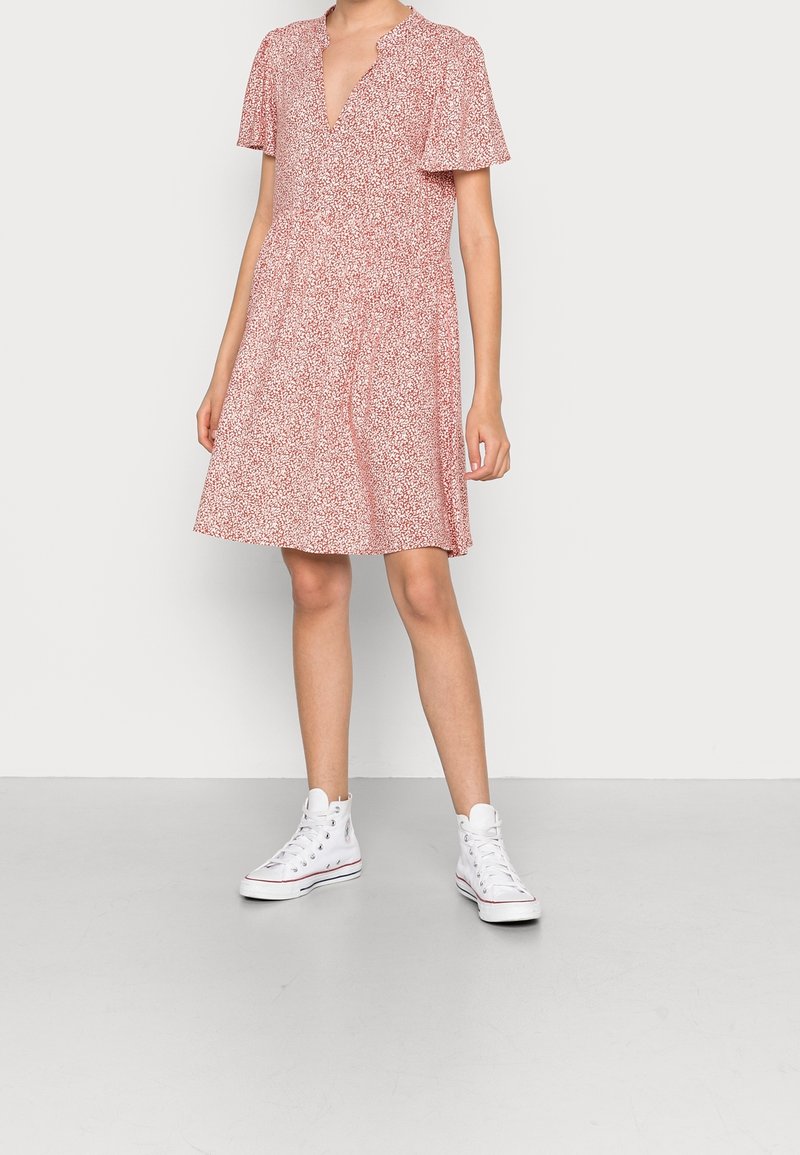 Femme portant une robe fleurie rose et blanche à manches courtes avec un col en V, associée à des baskets blanches montantes, debout sur un sol gris.