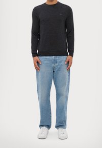 Muž v tmavě šedém svetru s dlouhým rukávem, světlých modrých džínách a bílých teniskách stojící na pozadí z čistě bílé barvy.