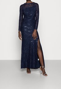 Navy lace evening gown with long sleeves, sequins, and a thigh-high slit. Worn with clear heeled sandals on a neutral background.