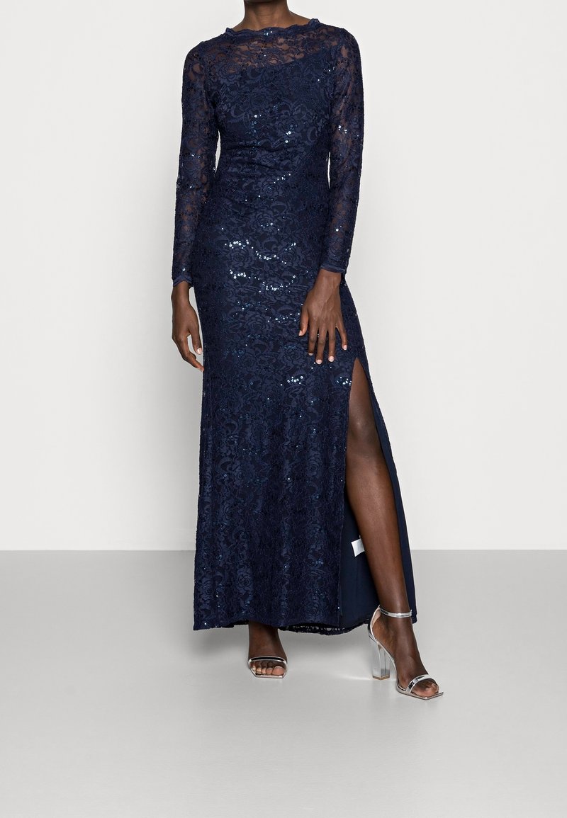 Navy lace evening gown with long sleeves, sequins, and a thigh-high slit. Worn with clear heeled sandals on a neutral background.