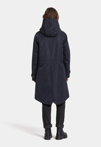 Marinblå vattentät jacka med huva, lång längd och dragsko i midjan. Har en enkel design med robusta detaljer och uppvikta ärmar.