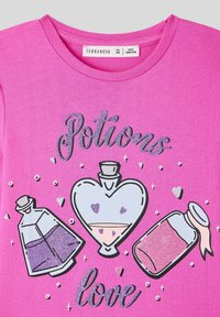 T-shirt rosa con design grafico di bottiglie di pozioni in varie forme. Accenti di glitter e cuori circondano il testo "L'amore per le pozioni".