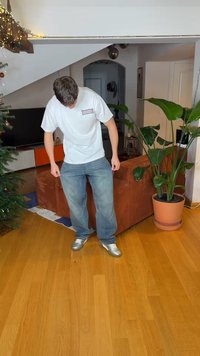 Młody mężczyzna w białej koszulce i dżinsach, patrzący w dół, stojąc na drewnianej podłodze w udekorowanym salonie z roślinami i choinką.