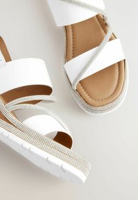 Sandales blanches avec des larges brides, semelle intérieure beige et épaisse semelle blanche. Décorées de strass argentés autour de la plateforme.