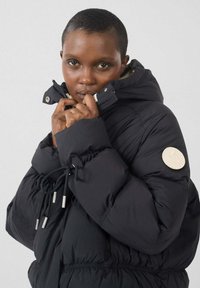 Zwarte pufferjas met een hoge kraag, elastische taillekoorden en een gestructureerde logopatch op de mouw. Voorzien van zilverkleurige hardware accenten.