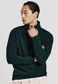 Dunkelgrüner, grob gestrickter Rollkragenpullover mit gerippten Bündchen und Saum. Das Modell hält die Hand nah am Hals, um Textur und Passform zu zeigen.