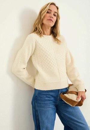 Knit cremefarbenes Sweatshirt mit strukturierten Mustern und geripptem Saum, kombiniert mit blauen Jeans. In einer Hand wird eine braun- und cremefarbene Umhängetasche gehalten.