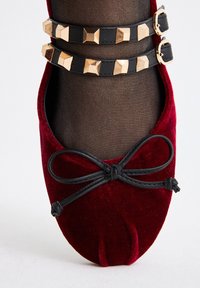 Bailarinas de terciopelo burdeos con una correa de cuero negro adornada con tachuelas en forma de pirámide doradas, y un detalle de lazada negra en la puntera.