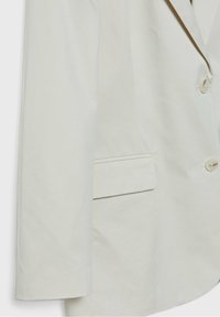 Manche de blazer écru avec poche à rabat et deux boutons visibles sur la fermeture avant, montrant une partie du revers.