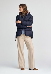 Granatowa kurtka puchowa z kapturem i sznurkiem, z zamkiem błyskawicznym i beżowymi akcentami, w połączeniu z kremowymi spodniami szerokimi i brązowym obuwiem.