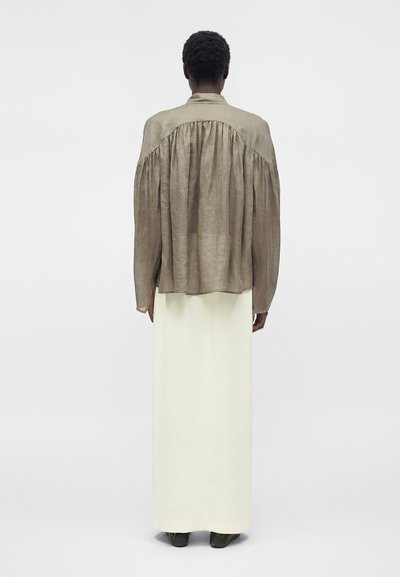 Άτομο με κοντά μαλλιά που φοράει ένα φαρδύ καφέ μακρυμάνικο πουκάμισο και μακρύ λευκό φούστα, στέκεται με την πλάτη σε απλό φόντο.